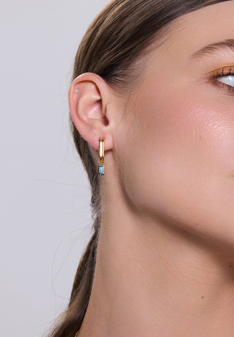 Złote kolczyki w kształcie obręczy o eleganckim wzornictwie z niebieskim prostokątnym kryształem. Gładka metalowa powierzchnia, minimalistyczny styl, lekkie.