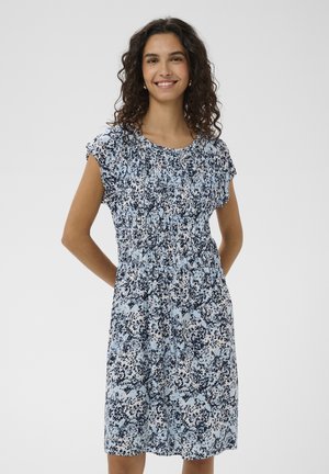 Žena s kučeravými tmavými vlasmi sa usmieva, má na sebe modro-čierne vzorované šaty s krátkym rukávom po kolená, stojí s rukami za chrbtom.