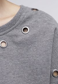 Sudadera gris con tela texturizada, con grandes ojetes metálicos en el hombro. Cuello redondeado, diseño informal y detalles de costuras visibles.