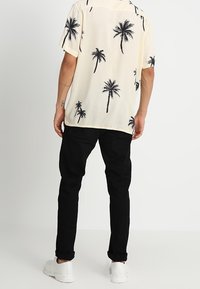 Camisa de manga corta en color crema con estampados de palmeras negras, combinada con pantalones negros y zapatillas blancas, vista desde atrás.