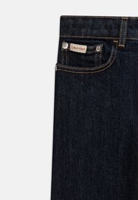 Jeans de denim oscuro con un botón plateado, costuras visibles en un color contrastante y una etiqueta que dice "Calvin Klein" en el bolsillo delantero.