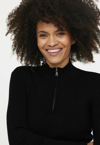 Schwarzer gerippter Pullover mit hohem Kragen und halbem Reißverschluss, der ein strukturiertes Design und eine taillierte Form aufweist, was für ein elegantes Erscheinungsbild sorgt.