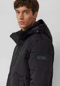 Zwarte pufferjas met een afneembare capuchon, voorzien van een doorgestikte textuur, een rits aan de voorkant, zijzakken en een gebrandmerkt label op de mouw.