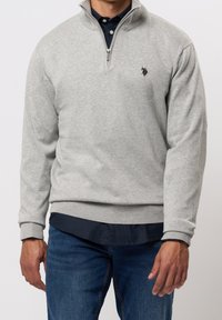 Grå zip-neck tröja med ribbade ärmslut och nederkant, lager på en mörk skjorta med knappar, har en broderad logotyp på bröstet.