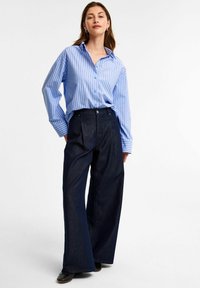 Niebiesko-biała, w paski koszula z guzikami o luźnym kroju, zestawiona z ciemnymi, szerokimi spodniami dżinsowymi. Outfit dopełniają czarne buty.