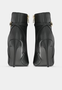 Botas de tobillo de cuero negro con tacones altos, con detalles de cremallera dorada y hebilla, vistas desde la parte posterior sobre un fondo blanco.