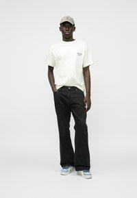 Witte korterm t-shirt met groene tekst, zwarte wijde jeans, grijze pet en blauwe en witte sneakers. Eenvoudige, casual outfit.