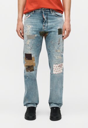 Homme portant un jean bleu clair déchiré avec plusieurs patchs en tissu et des baskets noires, debout devant un fond blanc.