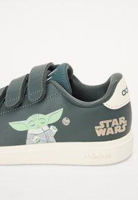 Vihreät synteettiset tennarit, joissa on kaksi tarranauhaa, ja joissa on painettu Baby Yodan grafiikka sekä kultainen "Star Wars"-logo sivussa.