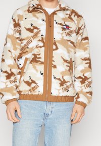 Giacca camouflage in peluche beige e marrone con cerniera color sabbia, polsini e vita; presenta una piccola etichetta di marca sul petto.