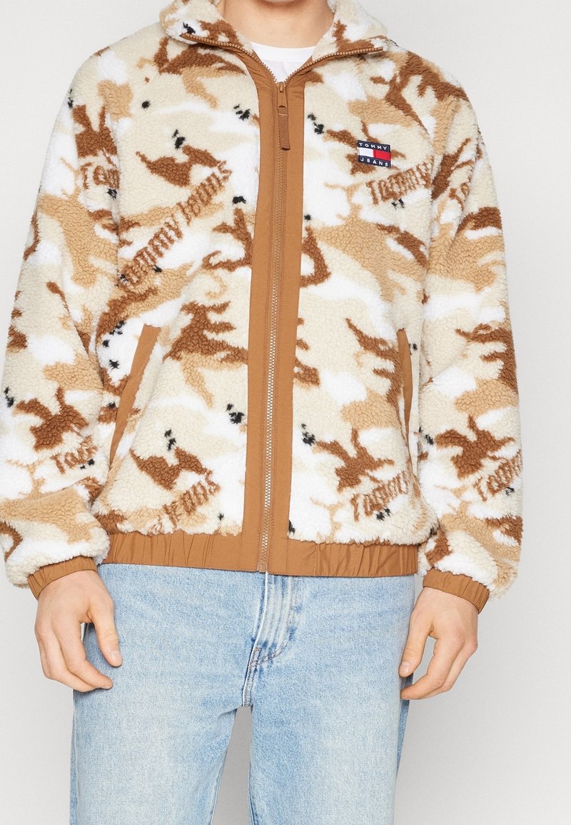 Giacca camouflage in peluche beige e marrone con cerniera color sabbia, polsini e vita; presenta una piccola etichetta di marca sul petto.
