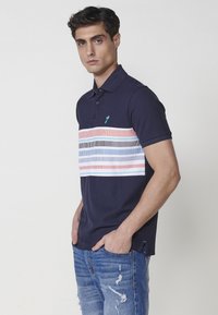 Tamsiai mėlynas polo marškinėliai su horizontaliomis juostelėmis rožinės, pilkos ir šviesiai mėlynos spalvų. Turi apykaklę ir subtilų logotipą ant krūtinės.