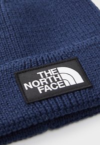 Marineblå strikket lue med en svart rektangulær lap med hvit "THE NORTH FACE" tekst og en kvart sirkel-logo på oppbretten.