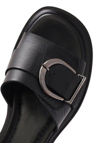 Sandalias negras con una banda de cuero ancha que presenta una hebilla de metal plateada. La suela es texturizada y el material superior parece suave.