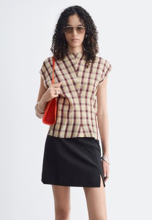 Femme aux cheveux bouclés porte un haut à carreaux marron, une jupe noire fendue, des lunettes de soleil, des bracelets en argent, des bagues et tient un petit sac à bandoulière rouge.