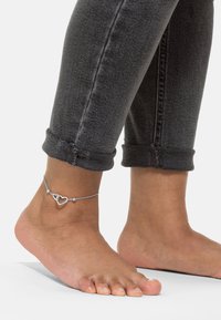 Sølv ankelarmbånd med to hjertecharms og små runde perler, båret på en bar fod, ved siden af grå optrillede jeans.