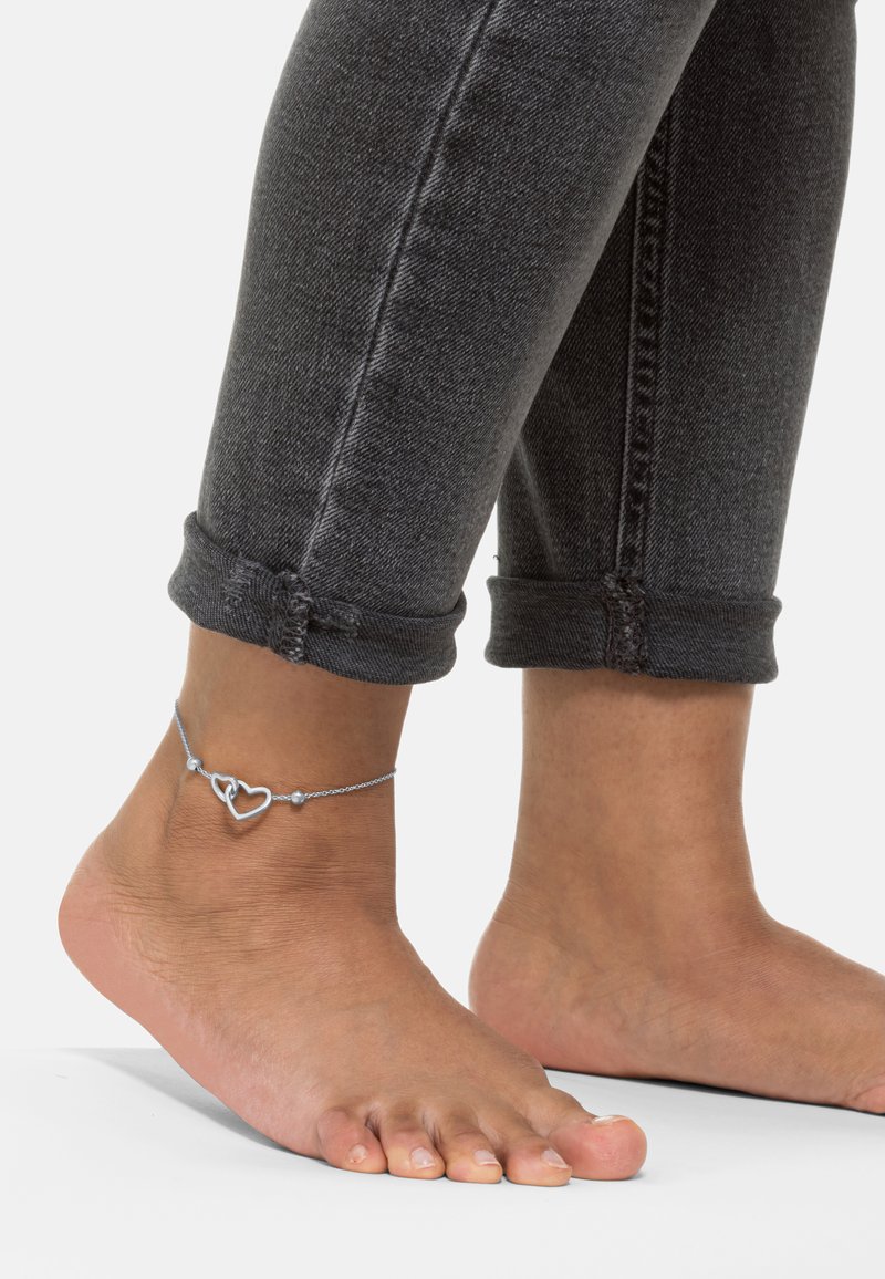 Sølv ankelarmbånd med to hjertecharms og små runde perler, båret på en bar fod, ved siden af grå optrillede jeans.