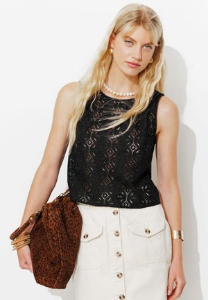 Femme blonde portant un top noir en dentelle sans manches, une jupe blanche boutonnée, des bijoux en or, et tenant un grand sac marron à imprimé léopard.