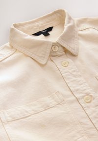 Camicia beige chiaro con bottoni, collo con risvolto, tessuto lavorato, tre bottoni e taschino sul petto sinistro. Etichetta del marchio visibile.