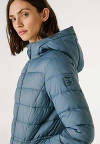 Vrouw met schouderlang bruin haar, gekleed in een lichtblauwe gewatteerde pufferjack met een capuchon, gezien vanaf de zijkant en halverwege de romp.