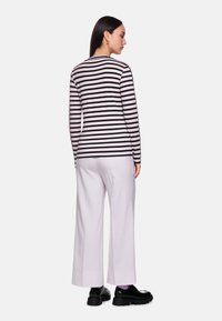 Frau, die sich abkehrt und ein schwarz-rosa gestreiftes Langarmshirt, hellrosa weite Hose und schwarze klobige Schuhe auf weißem Hintergrund trägt.