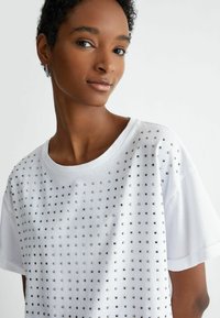 T-shirt in cotone bianco con una griglia di dettagli in borchie metalliche sul davanti, maniche corte e scollatura rotonda.