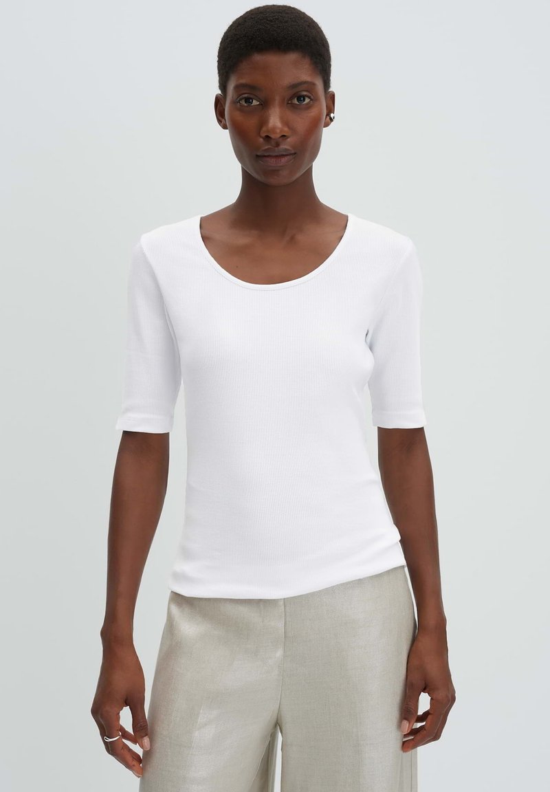 Vrouw met kort haar, gekleed in een wit aansluitend geribd shirt en lichtbeige wijde broek, staande tegen een effen grijze achtergrond.