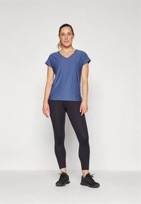 Mulher em pé vestindo uma blusa azul de mangas curtas e decote em V, leggings pretas e sapatilhas de desporto pretas, em um fundo branco liso.