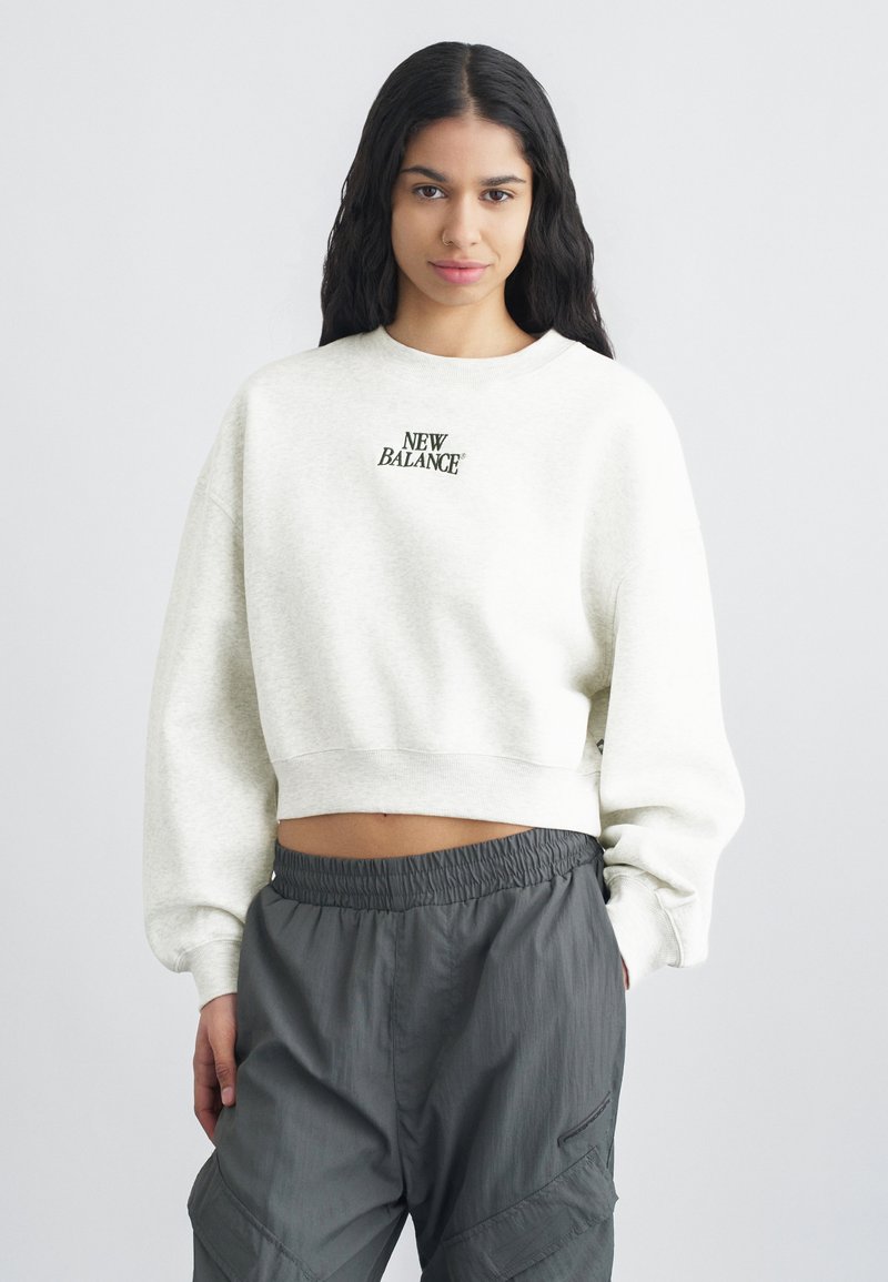 Νεαρή γυναίκα με μακριά μαύρα μαλλιά που φοράει λευκό κοντό φούτερ New Balance και χαλαρά σκούρα γκρι παντελόνια, στέκεται μπροστά σε απλό φόντο.