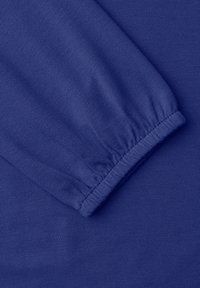 Blåt langærmet beklædningsstykke med elastisk manchet og synlig strikket tekstur, foldet for at vise ærmedetalje på matchende stofbaggrund.