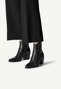 Bottines en cuir noir avec un bout pointu et un talon épais, dotées de panneaux élastiques sur les côtés et d'une texture lisse, associées à un pantalon noir large.