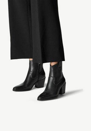 Bottines en cuir noir avec un bout pointu et un talon épais, dotées de panneaux élastiques sur les côtés et d'une texture lisse, associées à un pantalon noir large.
