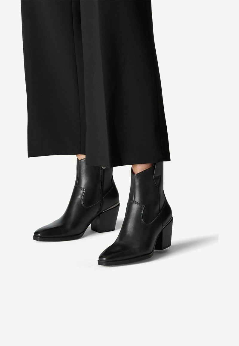 Bottines en cuir noir avec un bout pointu et un talon épais, dotées de panneaux élastiques sur les côtés et d'une texture lisse, associées à un pantalon noir large.