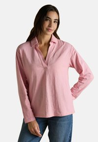 Naine, kellel on pikad tumedad juuksed, kannab heledat roosa värvi pika varrukaga polo särke ja siniseid teksaseid, seistes ühel käsivarrel puusal valge tausta ees.