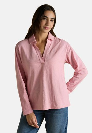 Naine, kellel on pikad tumedad juuksed, kannab heledat roosa värvi pika varrukaga polo särke ja siniseid teksaseid, seistes ühel käsivarrel puusal valge tausta ees.