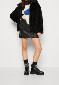 Schwarze Kunstfelljacke, gemusterter grauer Pullover mit blau-weißer Gestaltung, schwarzer Leder-Minirock und schwarze Schnürstiefel mit lila Akzenten.