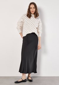Fleckiger Cream-Strickpullover mit gerippten Bündchen, kombiniert mit einem eleganten schwarzen Satin-Maxikleid und schwarzen flachen Schuhen. Das Outfit vereint verschiedene Texturen und Formen.