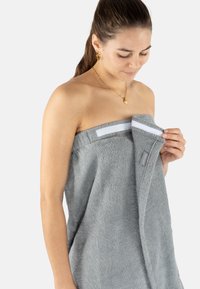 Grau Handtuchwickel aus strukturiertem Baumwollmaterial, mit einem horizontalen weißen Streifen und einem silbernen Etikett, das oben mit Klettverschluss befestigt ist.