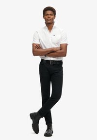 Camisa blanca de manga corta con un bolsillo en el pecho, combinada con pantalones ajustados negros y botas negras, con un diseño elegante y colores sólidos.