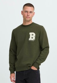 Ung mann med kort hår iført en olivengrønn hettegenser med et stort hvitt bokstav "B" på brystet, stående med en hånd i lommen.