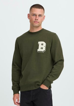 Young man with short hair wearing an olive green sweatshirt with a large white letter "B" on the chest, standing with one hand in pocket.