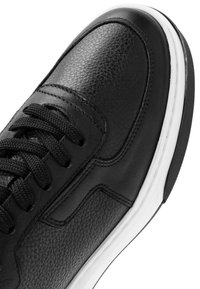 Close-up van een zwarte leren sneaker met textuur- en gladde panelen, zwarte veters en een witte zool met zichtbaar zwart profiel.