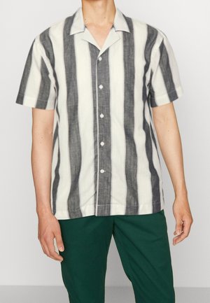 Mann trägt kurzärmeliges Hemd mit breiten vertikalen schwarzen und weißen Streifen und dunkelgrünen Hosen, steht vor einfarbigem Hintergrund.