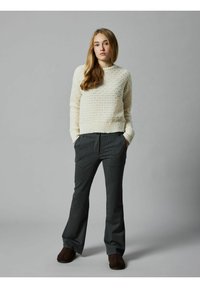 Helle Creme, strukturierter Strickpullover mit Rundhalsausschnitt; graue gestreifte Hose; braune Schlupfschuhe; das Modell steht mit den Händen in den Taschen.