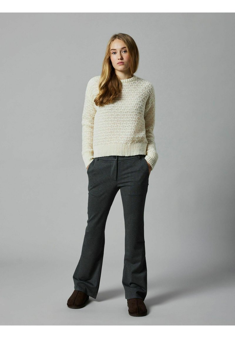 Helle Creme, strukturierter Strickpullover mit Rundhalsausschnitt; graue gestreifte Hose; braune Schlupfschuhe; das Modell steht mit den Händen in den Taschen.