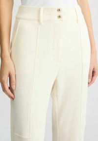 Pantaloni crema a vita alta con doppio bottone dorato in vita, tasche laterali e un tessuto liscio con vestibilità sartoriale.