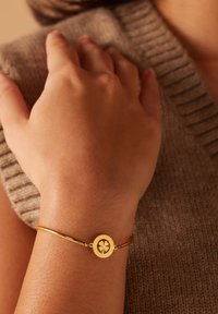 Gouden armband met een ronde hanger met een uitsnede van een klavertje. Gladde afwerking, gedragen op een hand tegen een getextureerde beige trui.