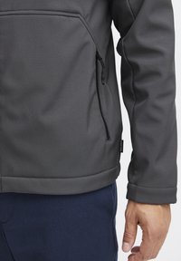 Grå softshell-jakke med en lynlåslomme og syning detaljer. Glat tekstur og buet kant, parret med marineblå bukser.