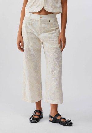 Primo piano di una persona che indossa pantaloni a gamba larga color crema con ricami floreali e sandali neri con borchie e fibbie argentate.