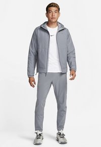 Viegls pelēks rāvējslēdzēja džemperis ar kapuci, saskaņots ar pelēkiem biksēm. Audums izskatās gluds ar sportiskām dizaina iezīmēm. Melns logotipa akcentu.